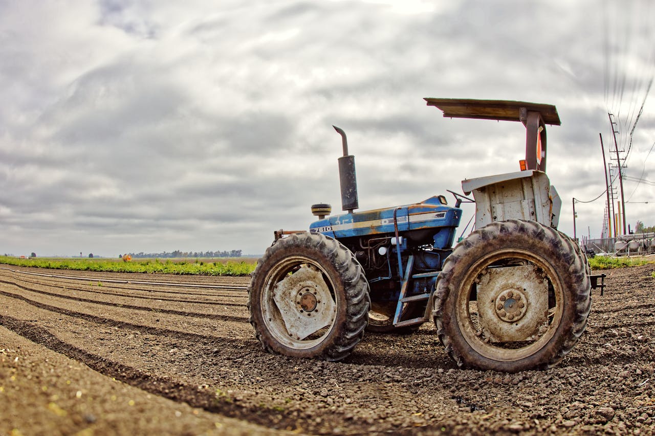 A vintage blue tractor sits idle on freshly plowed farmland under a cloudy sky.